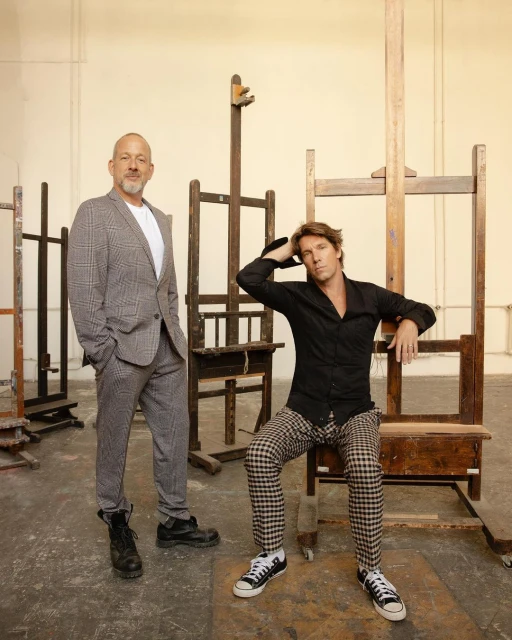 Two men pose in front of easels in a studio setting. One man wears a grey suit, and the other wears a black shirt and plaid pants.