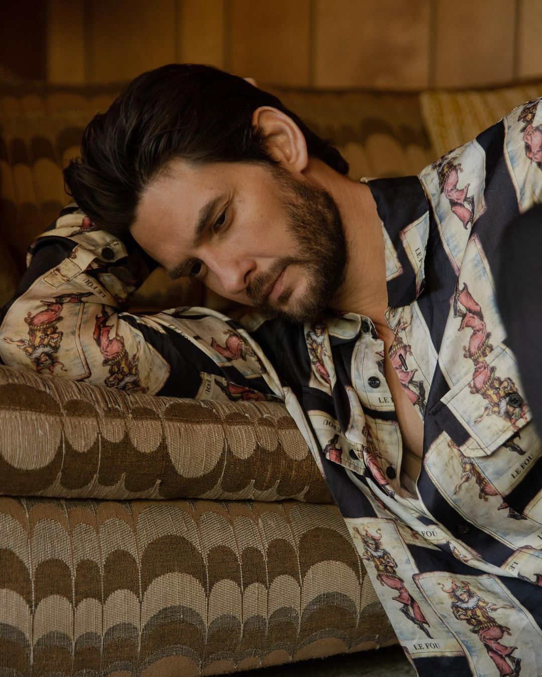 Adult male with dark hair lying on a patterned couch wearing a printed button-down shirt with a thoughtful expression and soft lighting.