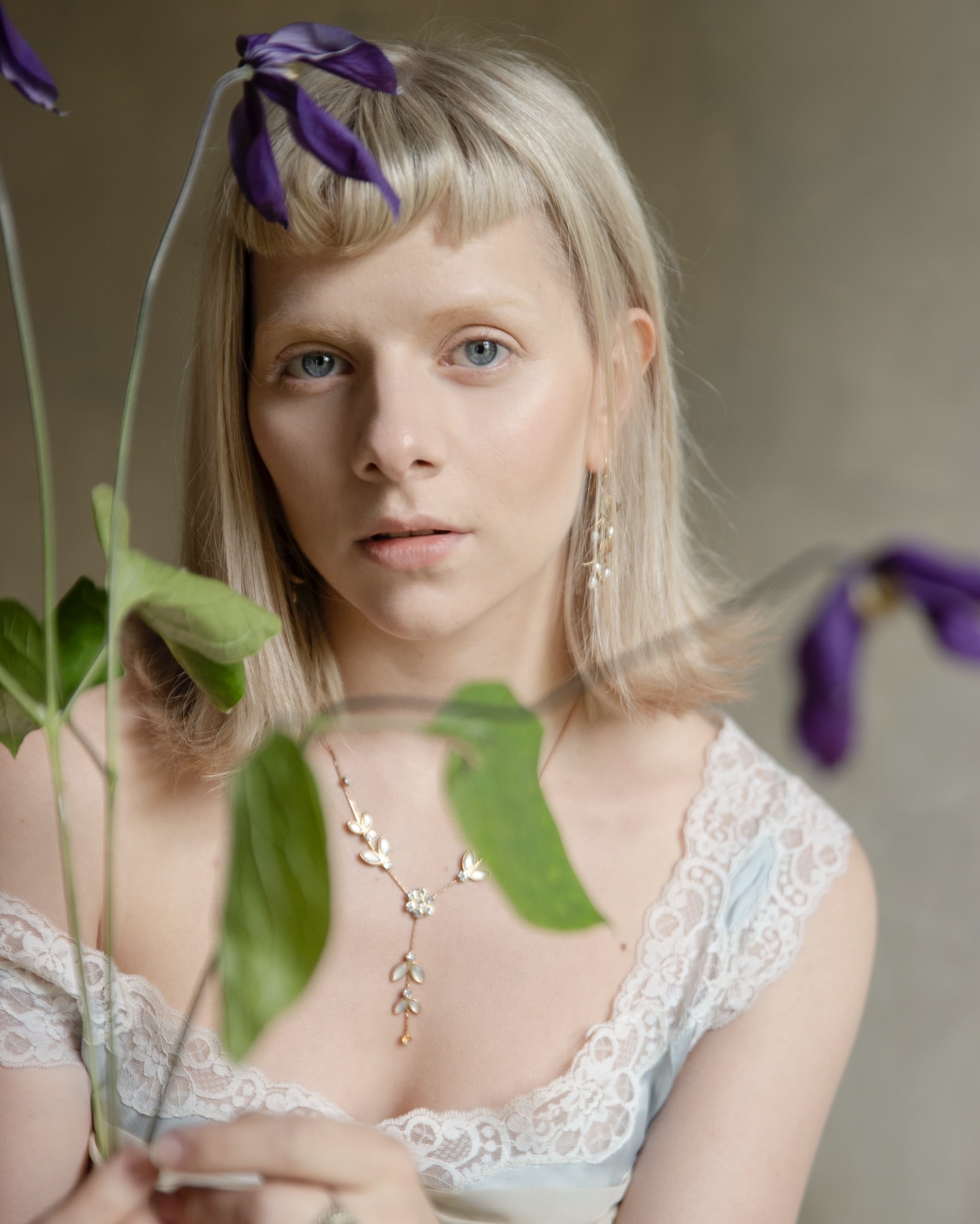 Close-up portrait of a young woman with blonde hair and blue eyes, wearing a lacy top and jewelry, holding purple flowers.