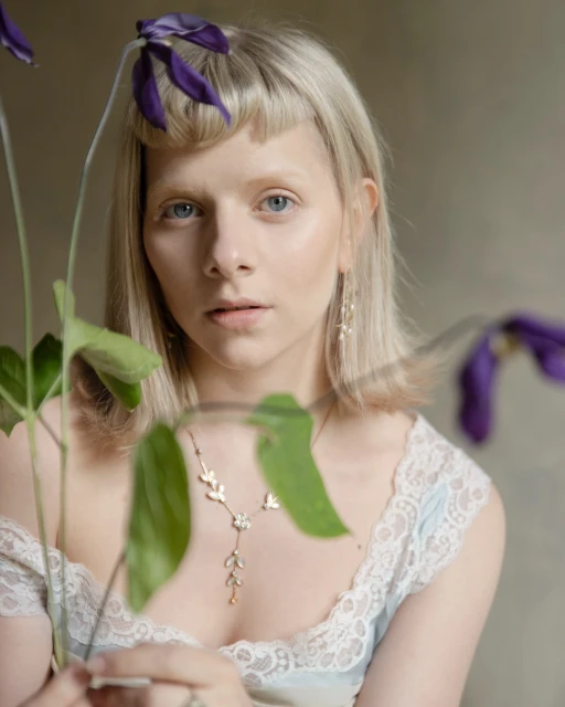 Close-up portrait of a young woman with blonde hair and blue eyes, wearing a lacy top and jewelry, holding purple flowers.