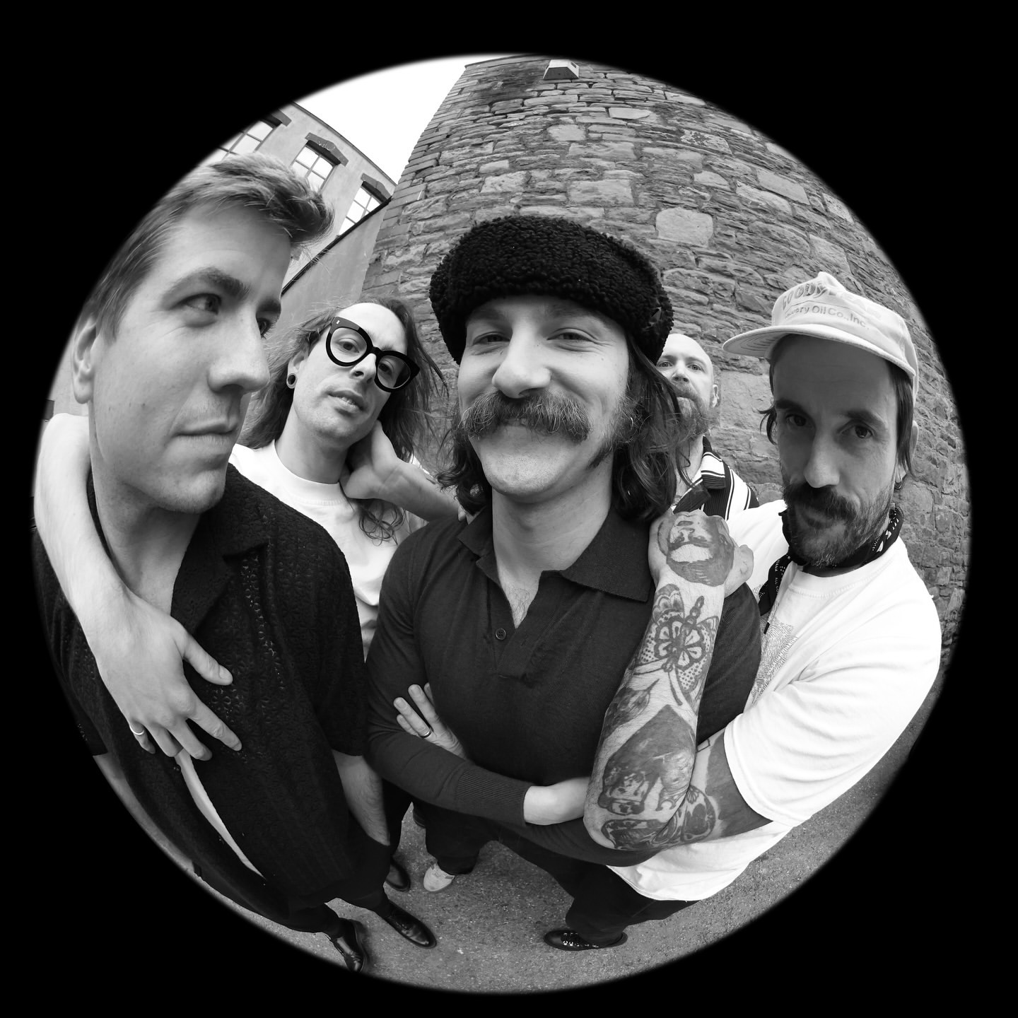Black and white fisheye view of a group of five men with facial hair posing in front of a stone building outdoors on a sunny day.