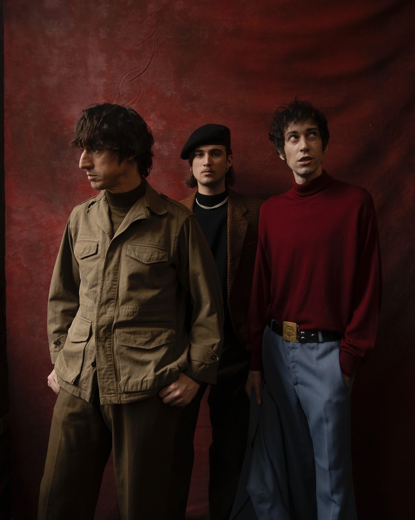 Three young adult males pose indoors against a red textured backdrop. One wears a beret, another a brown jacket and pants, one a red shirt.
