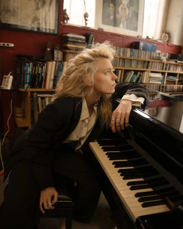 Blonde woman wearing a black blazer and white shirt seated at a piano in a room with a bookshelf background.
