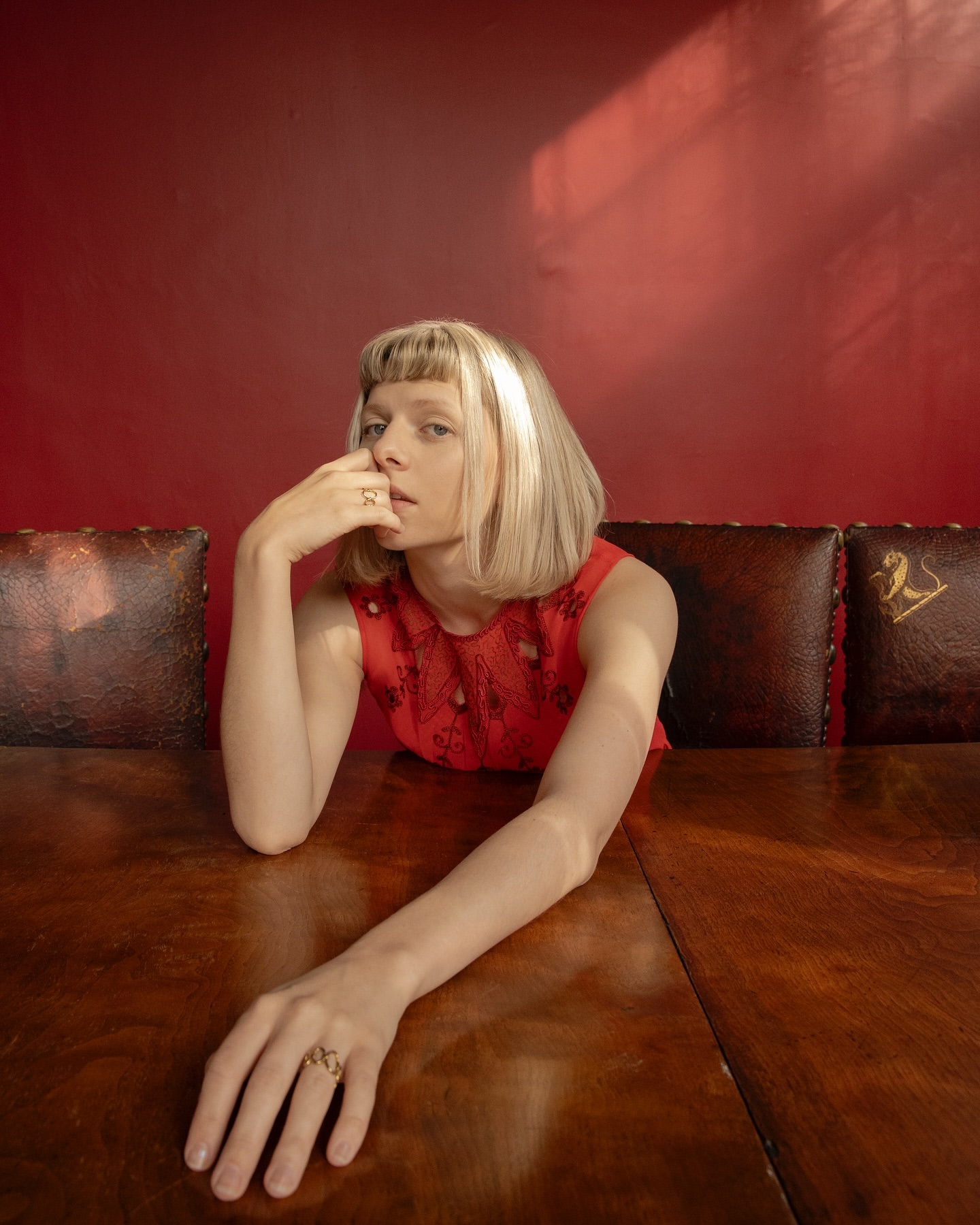 Portrait of a blonde woman with bangs in a red dress sitting at a wooden table against a red wall with shadow and decorative leather chairs.