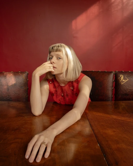 Portrait of a blonde woman with bangs in a red dress sitting at a wooden table against a red wall with shadow and decorative leather chairs.