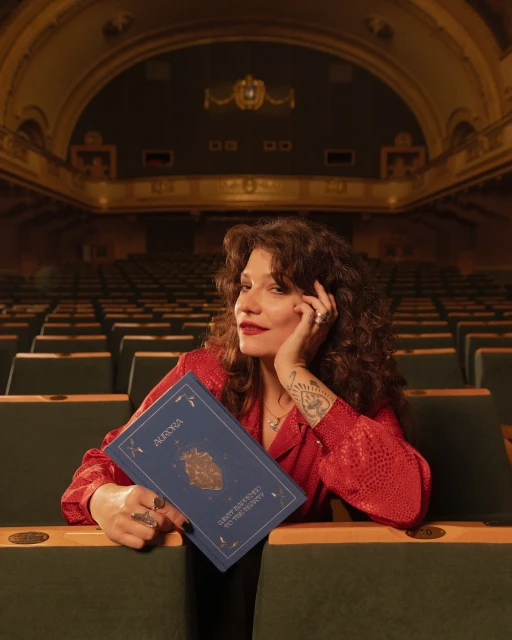 Woman in a red patterned shirt in a theatre holds a blue book titled AURORA, with empty seats filling the background.