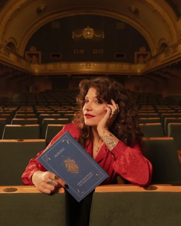 Woman in a red patterned shirt in a theatre holds a blue book titled AURORA, with empty seats filling the background.