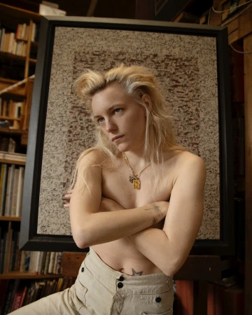Portrait of a blonde woman with arms crossed wearing beige trousers in front of a framed textured artwork.