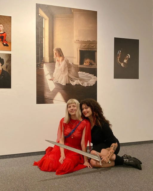 Two women sit on the floor of an art gallery holding a sword in front of posters featuring paintings on the wall behind them.