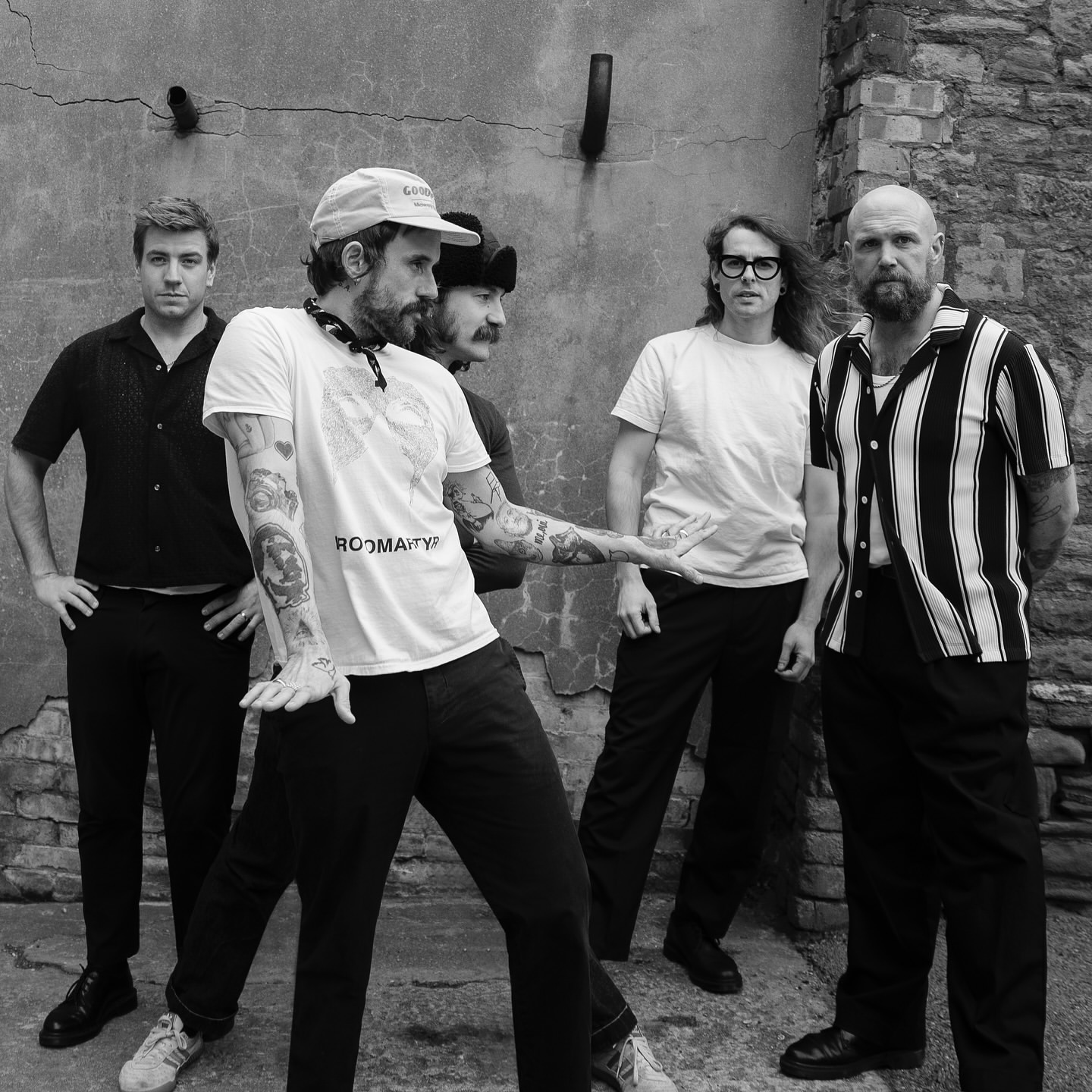 Five men stand outside a distressed stone building in a black and white portrait, wearing casual clothes and various facial hair styles.