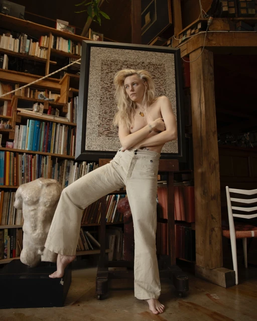 Bare-chested woman in wide-leg pants poses in a rustic, book-lined room with a framed artwork and a classical torso sculpture.