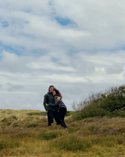 A couple embraces in a grassy field under a cloudy sky. The man wears a dark jacket and the woman a flannel shirt as they hug.