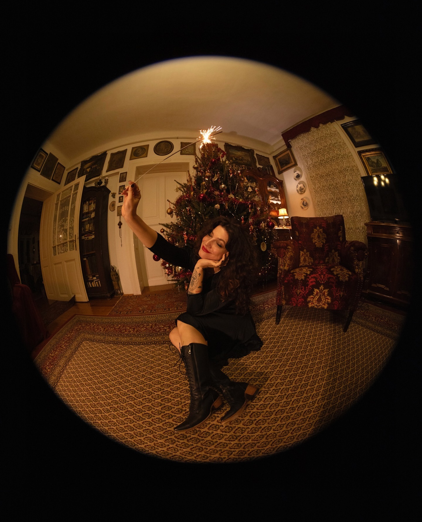 Woman in black dress and boots sits on floor near Christmas tree holding sparkler in fisheye view, creating a festive, distorted perspective.