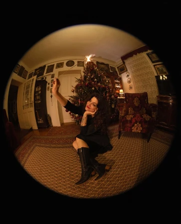 Woman in black dress and boots sits on floor near Christmas tree holding sparkler in fisheye view, creating a festive, distorted perspective.