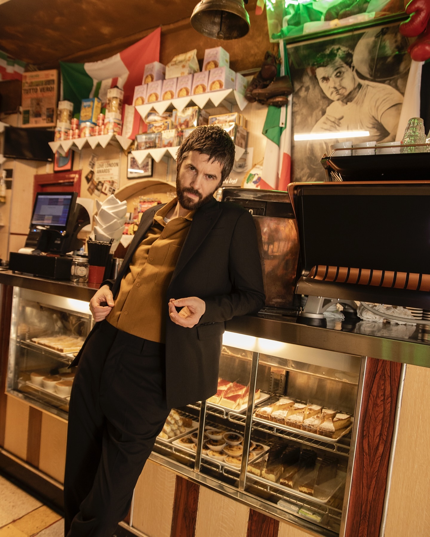 Man in suit leans on a deli counter with baked goods. Italian flags and a portrait hang in the background in a warm, indoor setting.