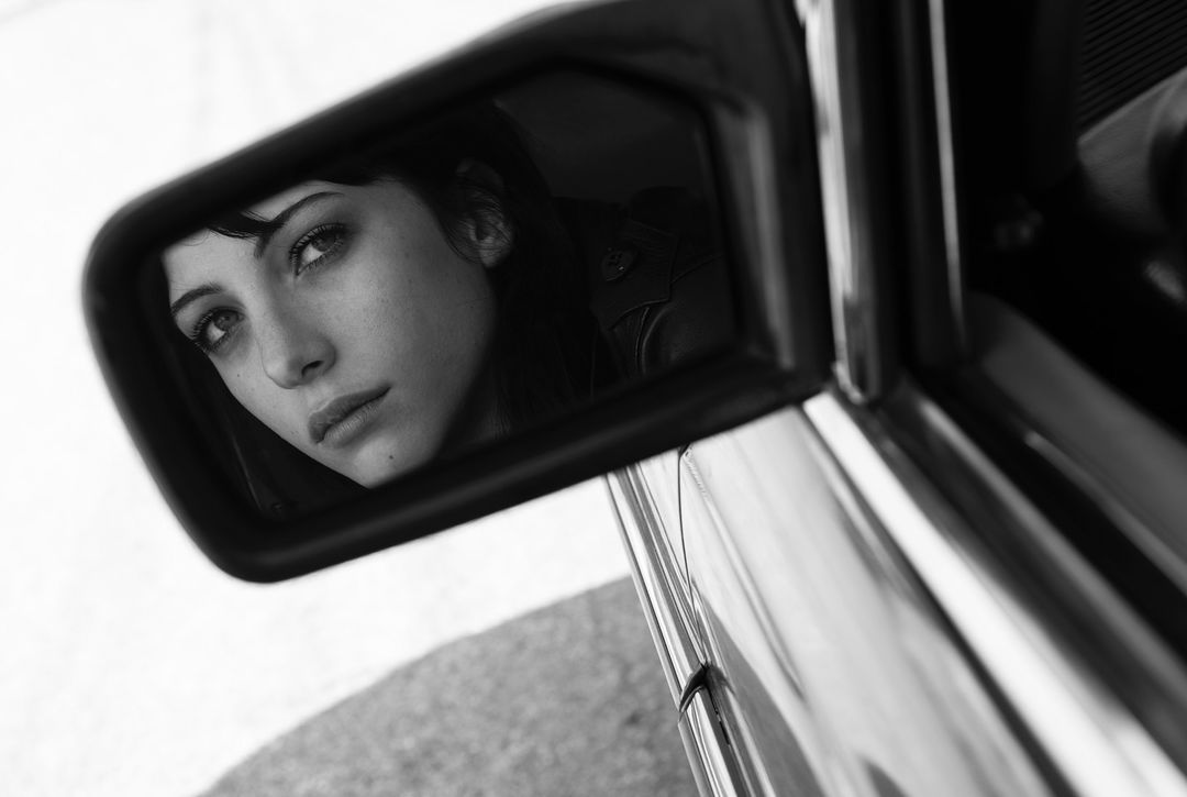 Black and white portrait of an adult female reflecting in a car side mirror, looking slightly upward with neutral expression and dark hair.