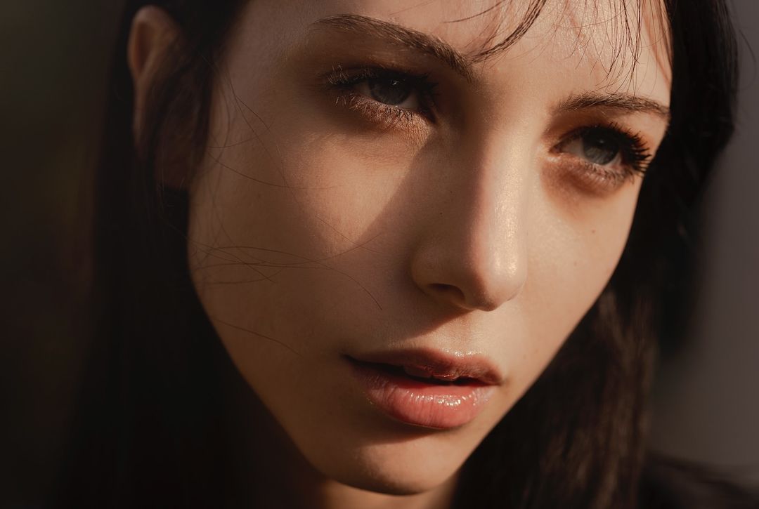 Close-up portrait of a young adult woman with natural light highlighting her features, set against a blurred, dark background.