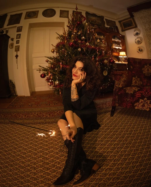 Woman in black dress and boots sits near decorated Christmas tree indoors with warm lighting, holding sparklers, creating a festive atmosphere.