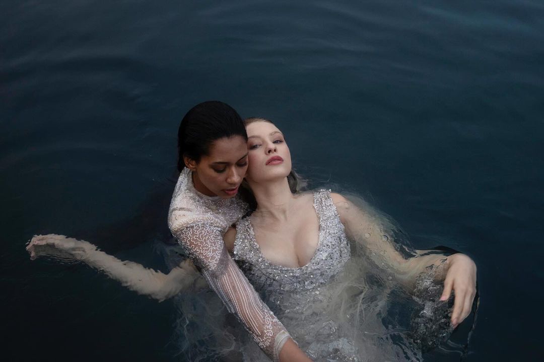 Two women in embellished gowns are submerged in water, one resting against the other, creating an intimate and artistic fashion image.