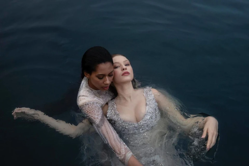 Two women in embellished gowns are submerged in water, one resting against the other, creating an intimate and artistic fashion image.