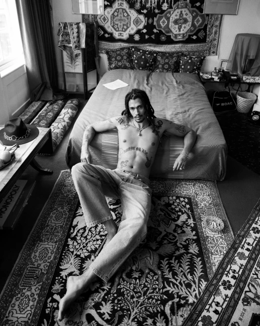 Black and white portrait of a man with tattoos lying on a patterned rug in a bedroom with a bed and various textiles.
