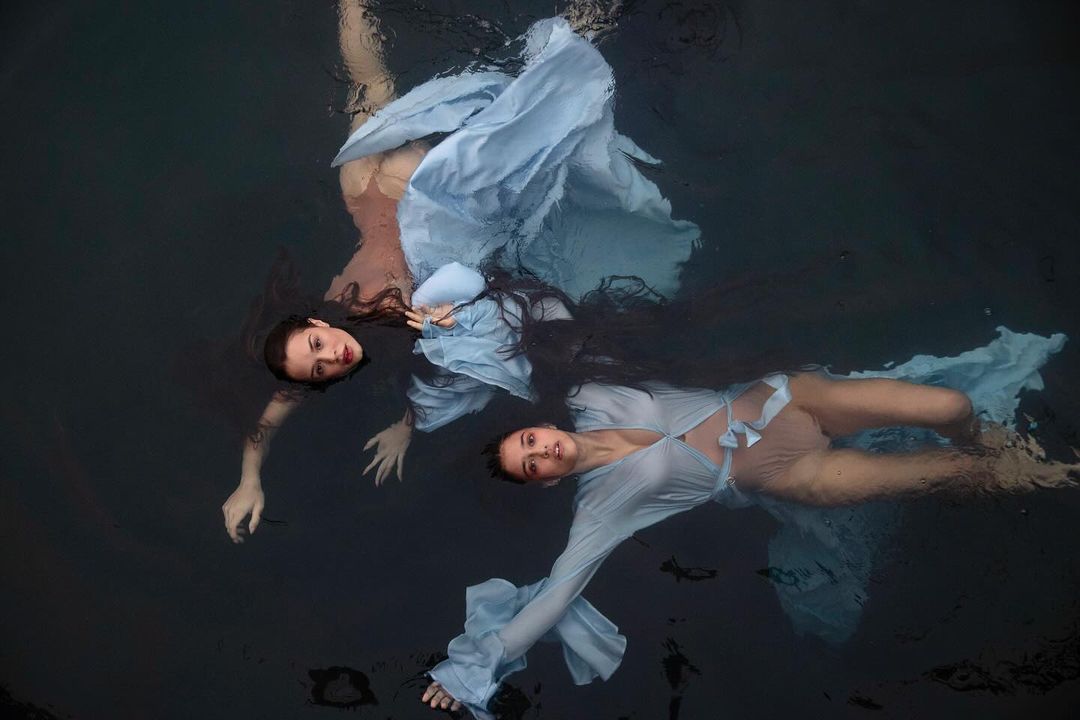 Two women in blue dresses float in dark water, creating a mirrored effect with reflections and dramatic, artistic underwater photography.