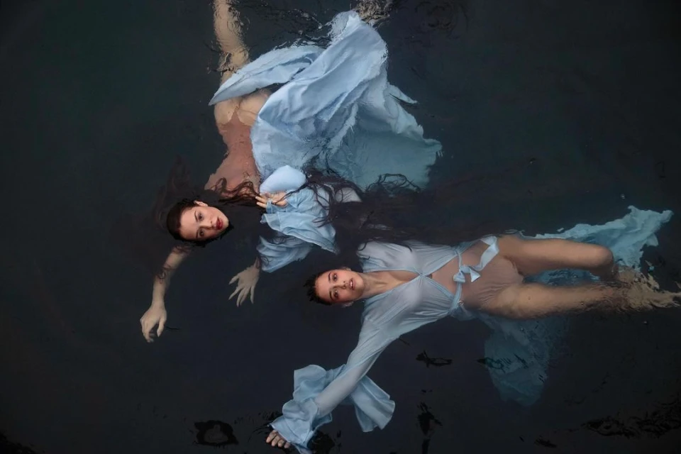 Two women in blue dresses float in dark water, creating a mirrored effect with reflections and dramatic, artistic underwater photography.