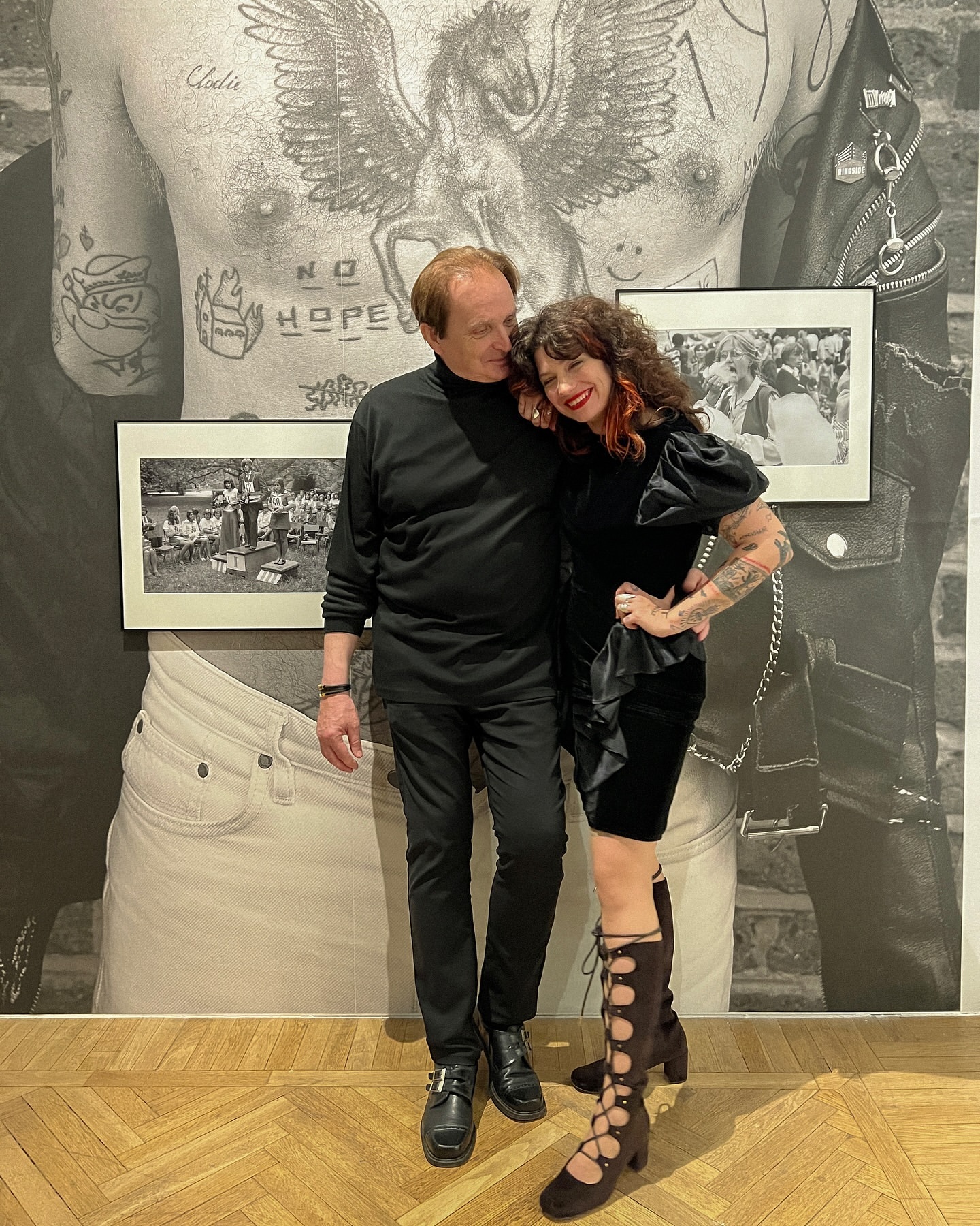 A couple poses indoors in front of large black and white photos. The woman wears a black dress and gladiator-style heels and the man wears all black.