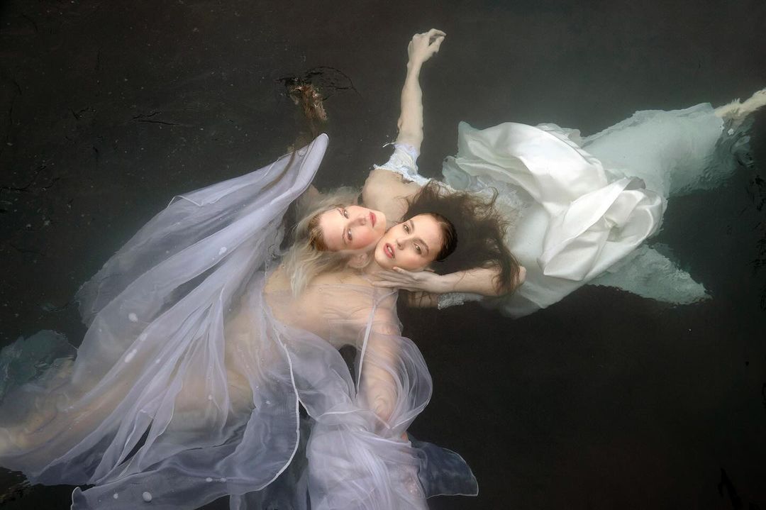 Two women are floating in dark water wearing light-colored dresses, hair flowing around them, creating a surreal underwater scene.