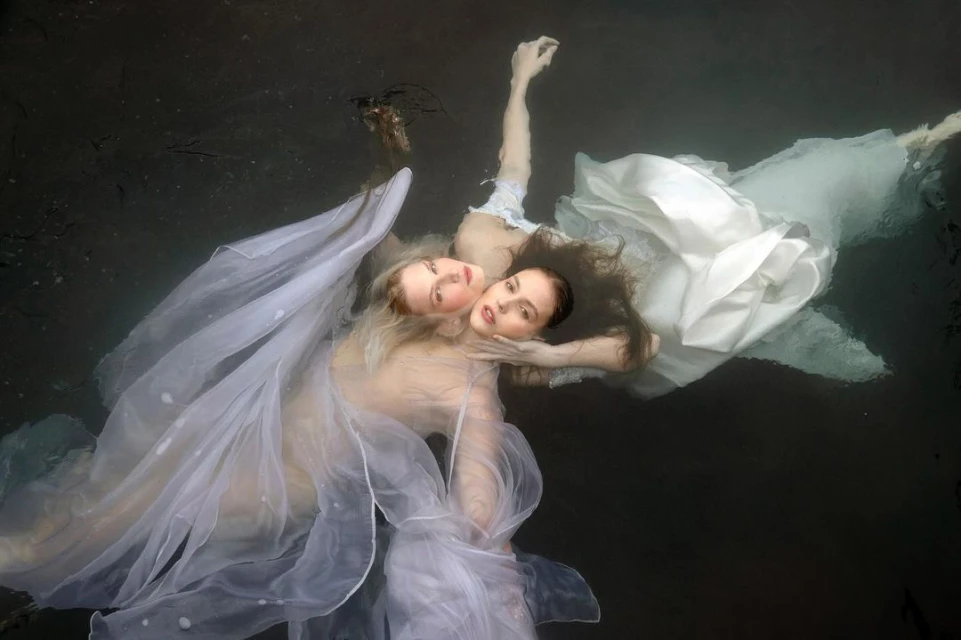 Two women are floating in dark water wearing light-colored dresses, hair flowing around them, creating a surreal underwater scene.