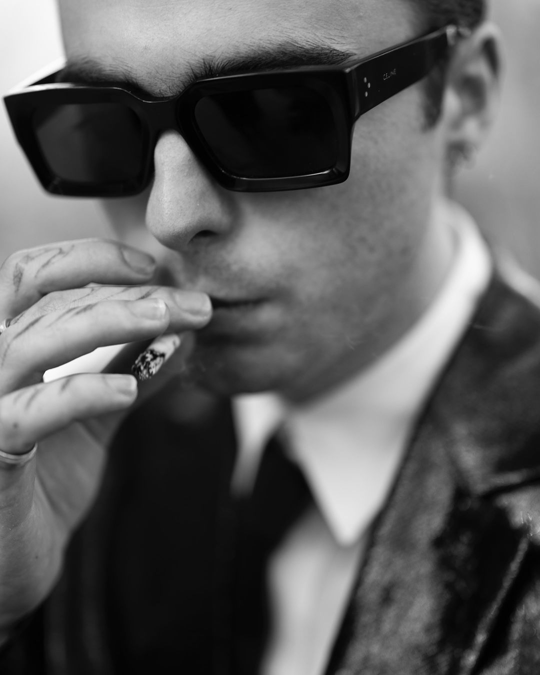 Close-up portrait of a man wearing thick black sunglasses and smoking a cigarette, photographed in London by Wanda Martin.