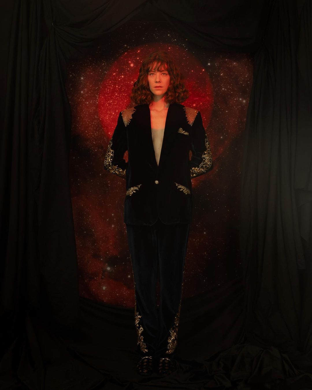 Adult woman in a black velvet suit with metallic embroidery posing in a studio with a red nebula and black draping.