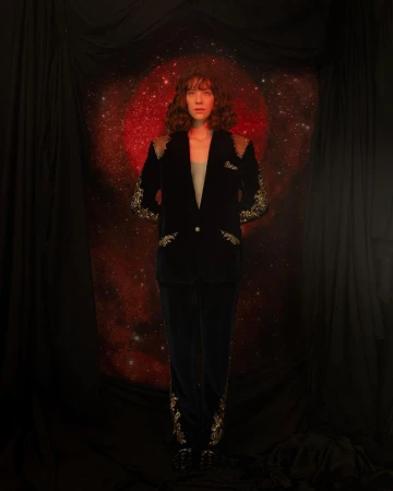 Adult woman in a black velvet suit with metallic embroidery posing in a studio with a red nebula and black draping.