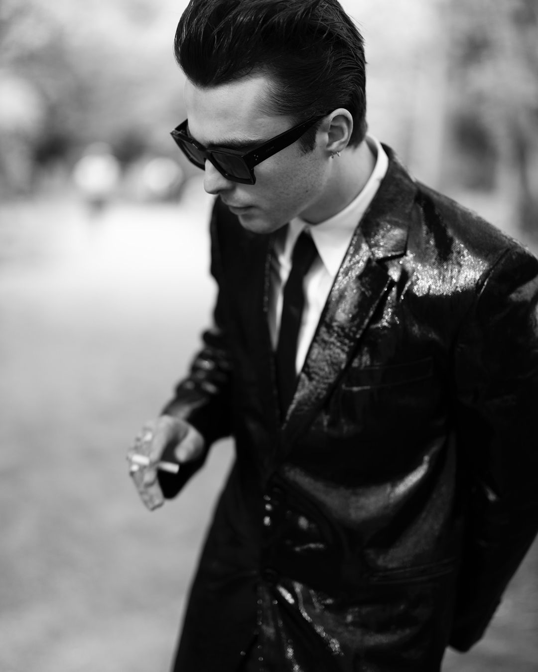 Man wearing sunglasses and a dark suit jacket holding a cigarette in black and white photography by Wanda Martin.