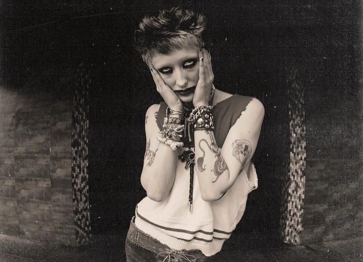 Portrait of a person with short hair, heavy eye makeup, tattoos, and layered bracelets holding their face in front of a dark tiled wall.