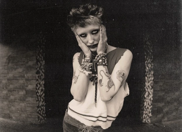 Portrait of a person with short hair, heavy eye makeup, tattoos, and layered bracelets holding their face in front of a dark tiled wall.
