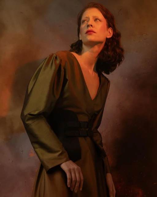 Portrait of a curly-haired woman with fair skin, wearing a dark olive green dress, looking up against a soft orange and brown background.