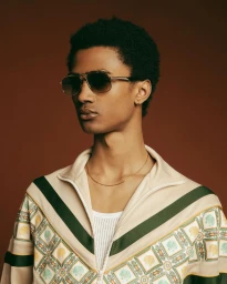 Portrait of a Black man wearing aviator sunglasses and a patterned zip jacket against a warm solid background