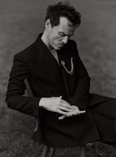 Monochrome portrait of a man in a black suit seated in a grassy outdoor setting, showcasing a classic, elegant, and contemplative pose.