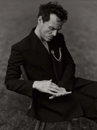 Monochrome portrait of a man in a black suit seated in a grassy outdoor setting, showcasing a classic, elegant, and contemplative pose.