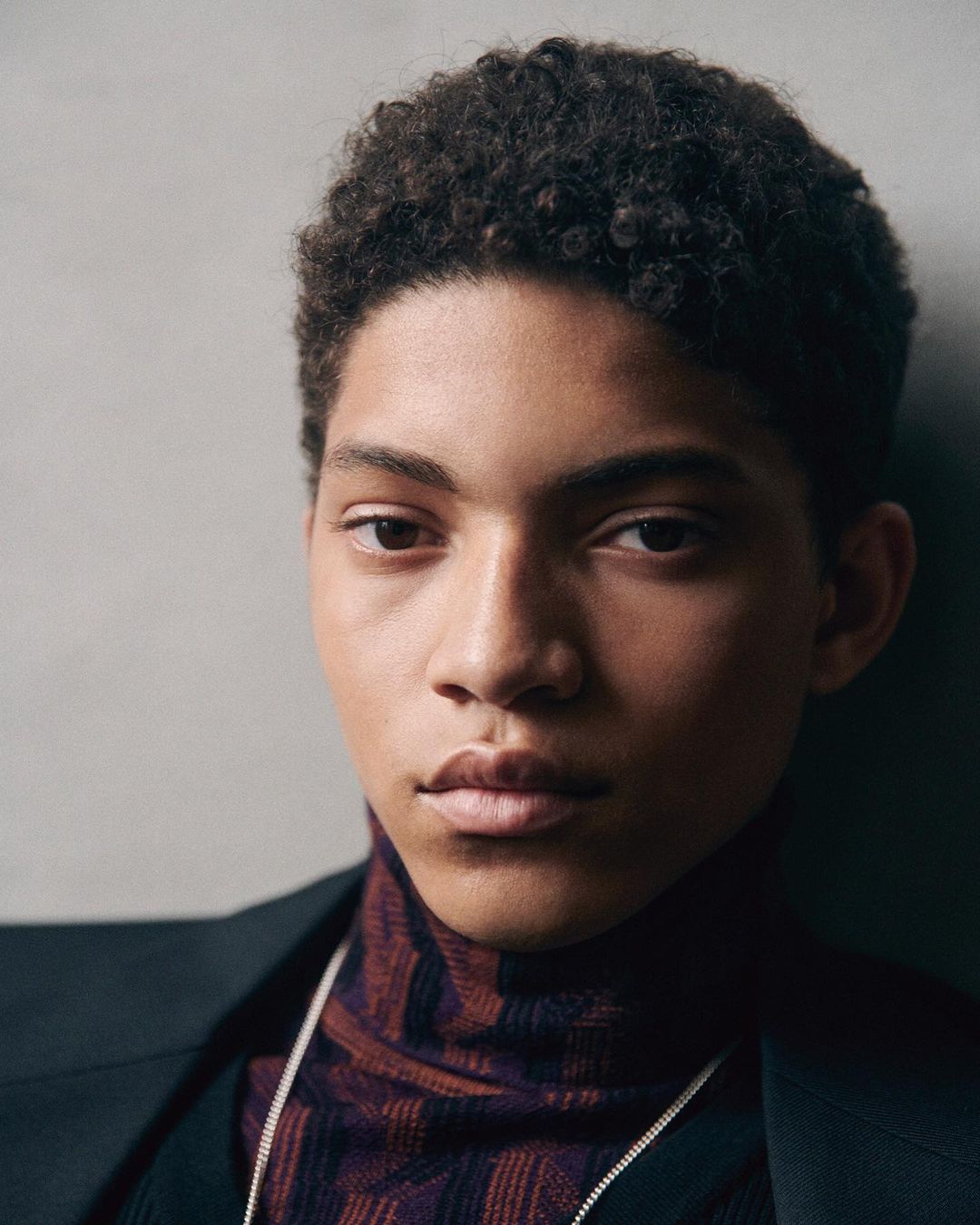 Close-up studio portrait of a young adult with dark curly hair wearing a patterned turtleneck and suit jacket against a grey background.