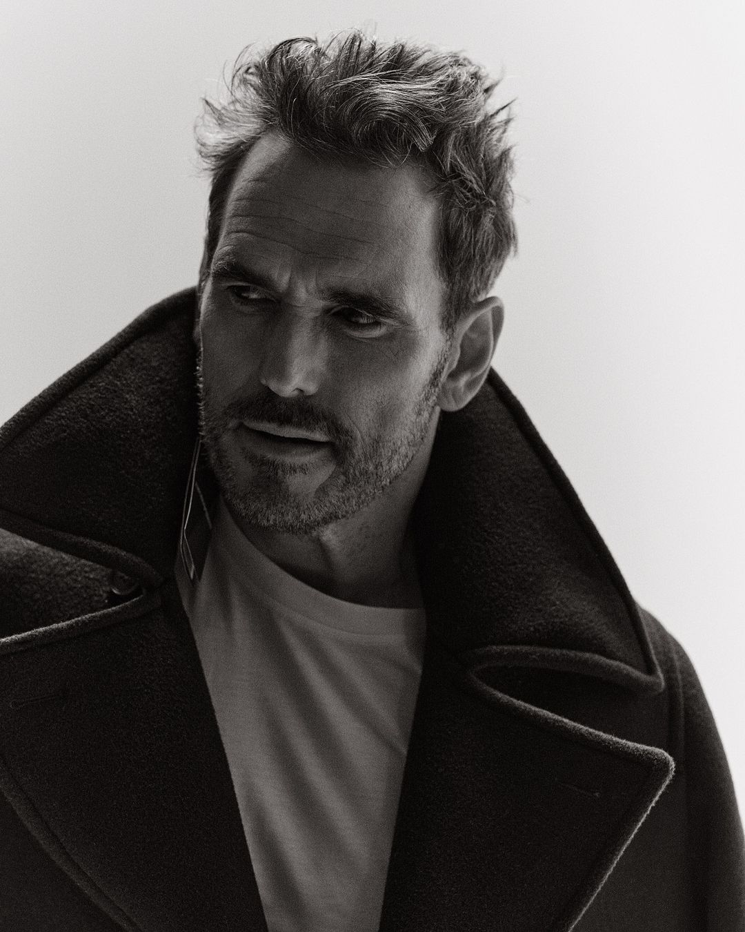 Monochrome portrait of a handsome adult male in a pea coat, thoughtfully gazing to the side, set against a bright white backdrop.
