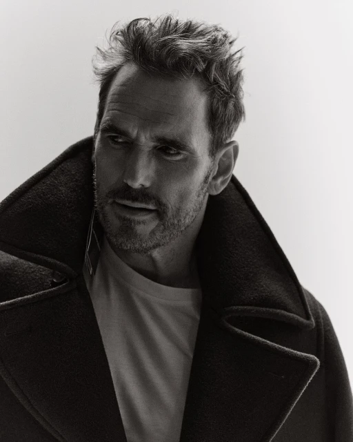 Monochrome portrait of a handsome adult male in a pea coat, thoughtfully gazing to the side, set against a bright white backdrop.