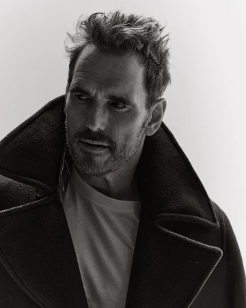 Monochrome portrait of a handsome adult male in a pea coat, thoughtfully gazing to the side, set against a bright white backdrop.