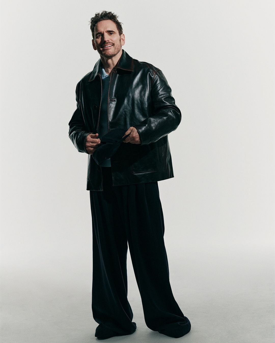 Matt Dillon wearing a dark leather jacket and wide trousers against a light grey background, photographed by Ward Ivan Rafik.