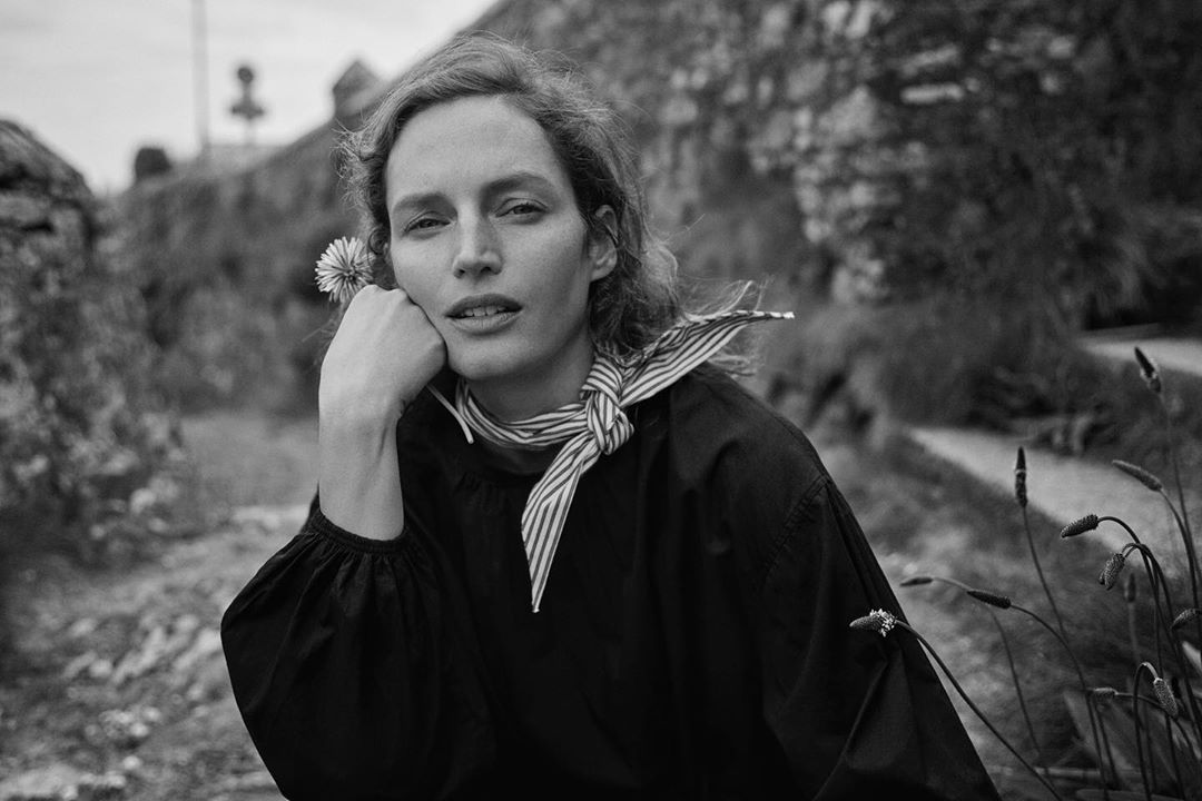 Black and white portrait of a young woman in a black top holding a flower in a rural environment, serene expression.