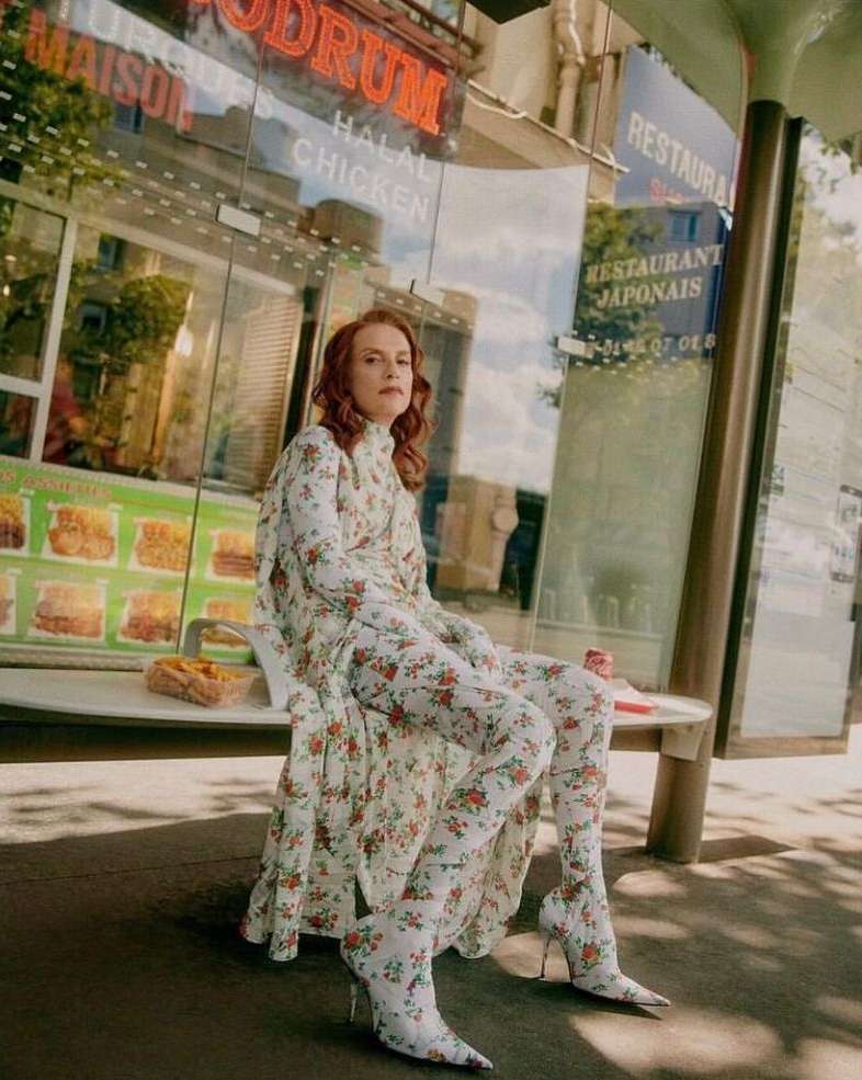 A woman in a floral dress and matching thigh-high boots sits outside of a restaurant in the city on a sunny day.
