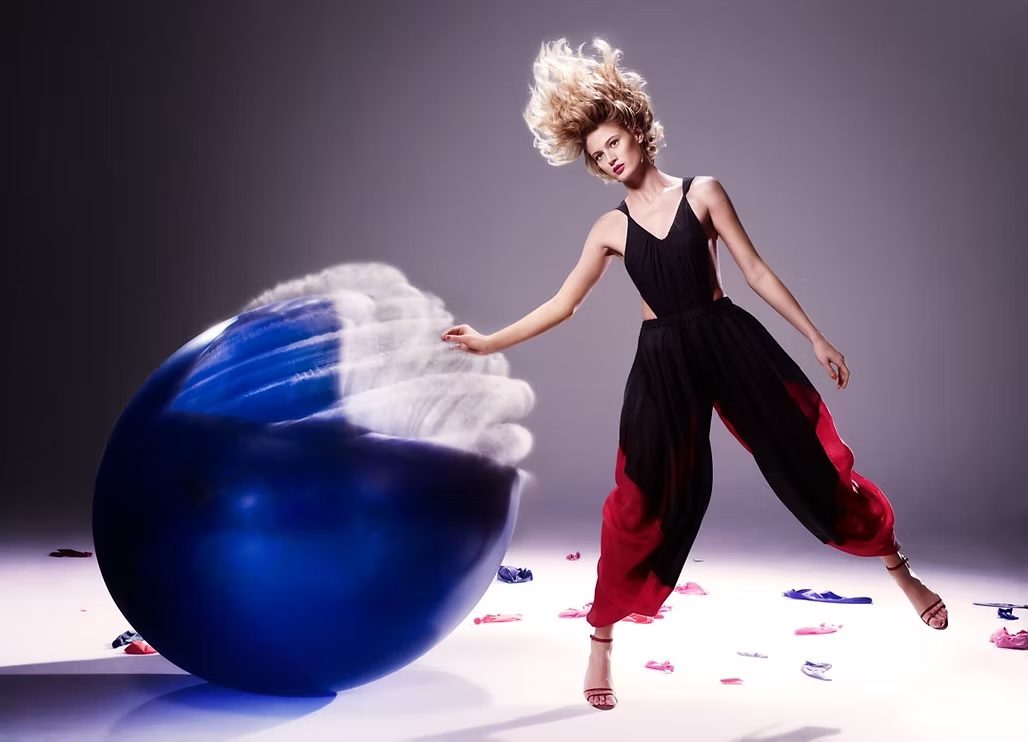 Fashion model in black and red jumpsuit strikes dynamic pose next to an oversized blue orb in a photo studio setting with scattered sandals.