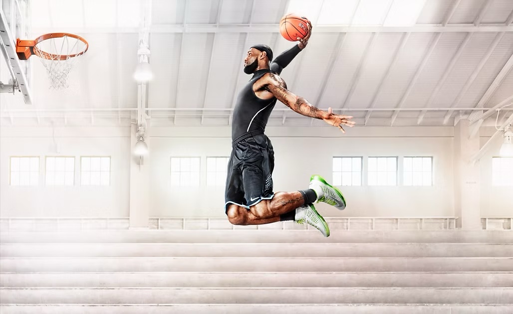 Athlete in mid-air, poised to dunk a basketball, wearing athletic gear against a bright, white gymnasium backdrop.