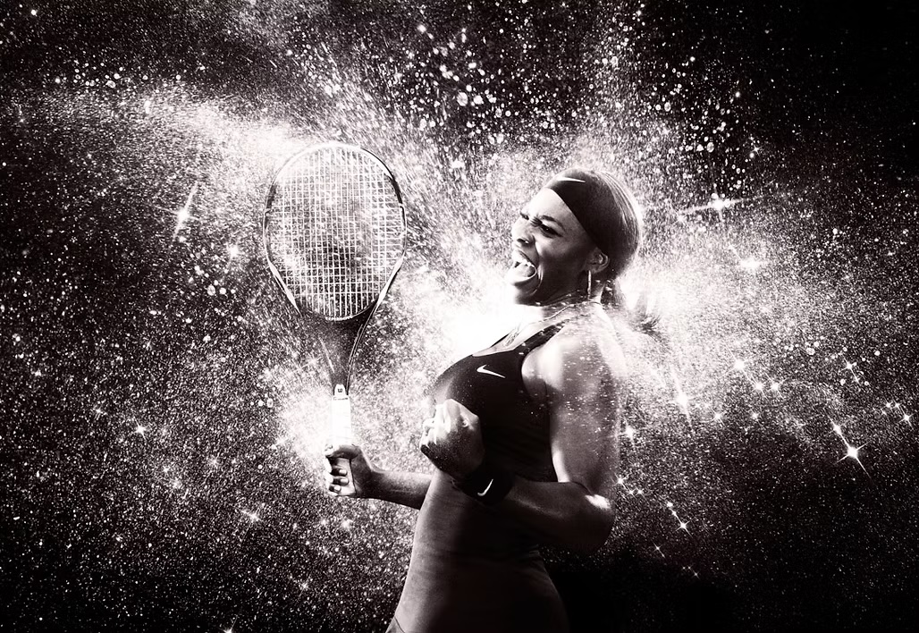 A monochromatic action shot of an adult woman celebrating with a tennis racket against a dark background, surrounded by water splash effect.
